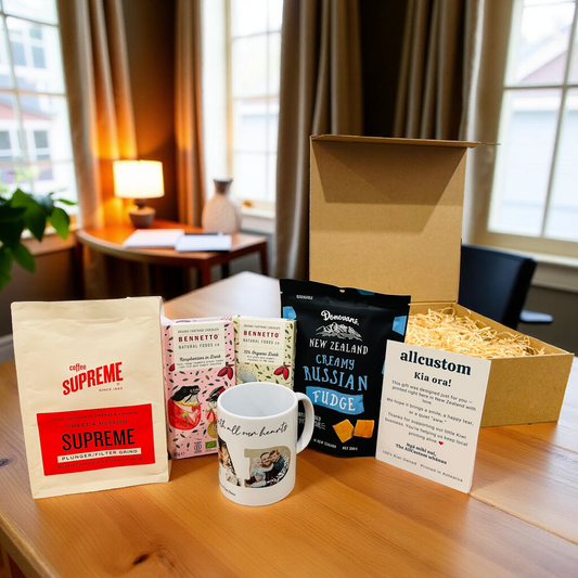 Gift box with coffee bag, mugs, and fudge on a wooden surface with a window in the background.