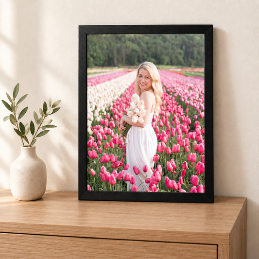 A4 framed photo print of a woman holding tulips in a flower field, black frame, displayed on a wooden sideboard next to a small vase