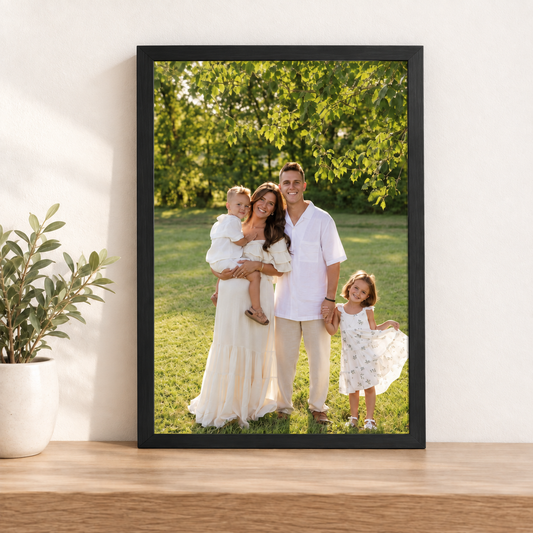 Framed photo print of a family of four in a park, A3 size in a black frame, standing on a wooden shelf beside a potted plant